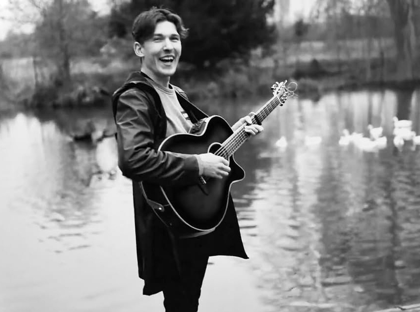 Michael Blackman playing acoustic guitar and smiling by a lake